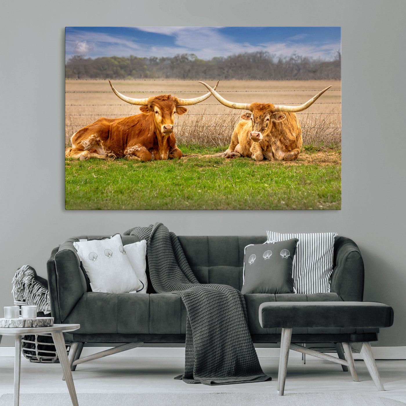 Two longhorn cows lying on green grass in front of a golden field and trees under a blue sky, perfect for rustic country wall decor.
