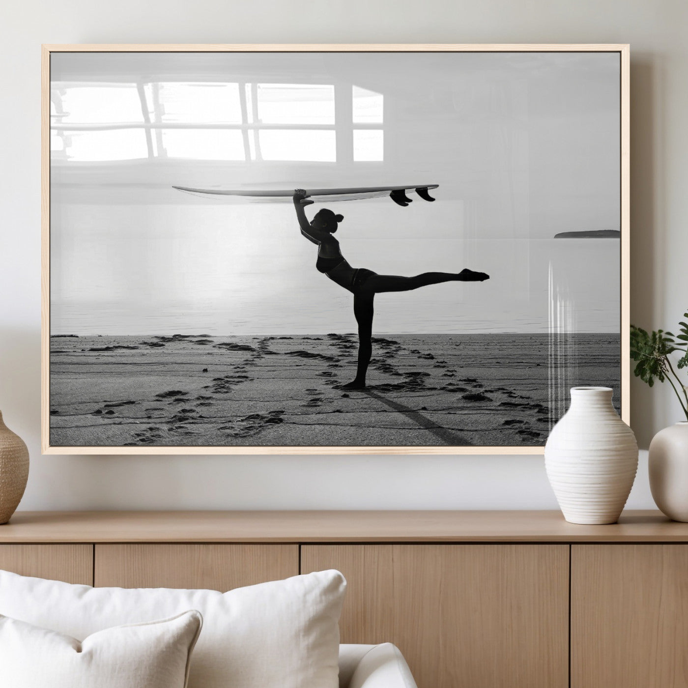 A woman balances a surfboard on her head while striking a yoga pose on the beach, silhouetted against a calm ocean horizon.