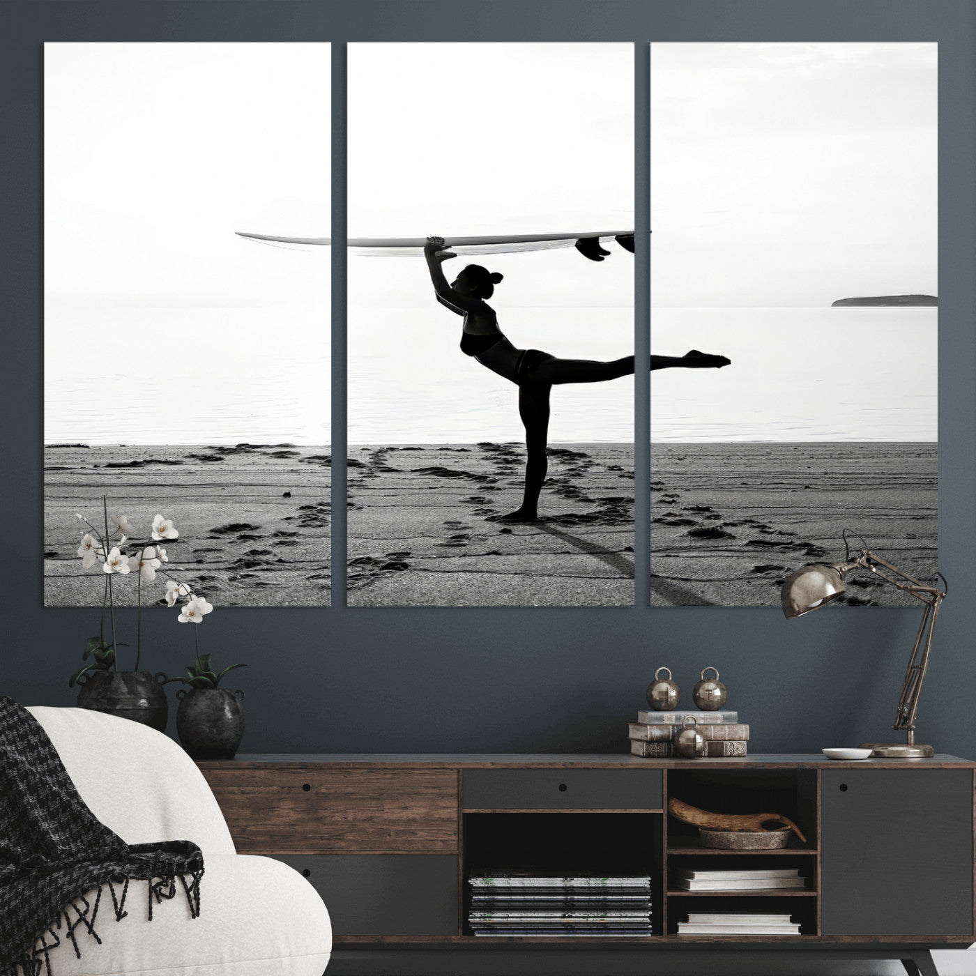 A woman balances a surfboard on her head while striking a yoga pose on the beach, silhouetted against a calm ocean horizon.