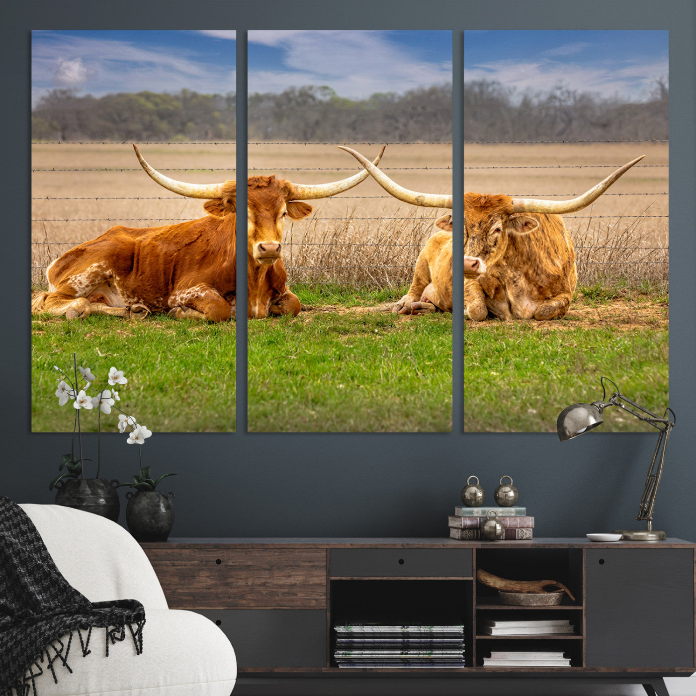 Two longhorn cows lying on green grass in front of a golden field and trees under a blue sky, perfect for rustic country wall decor.