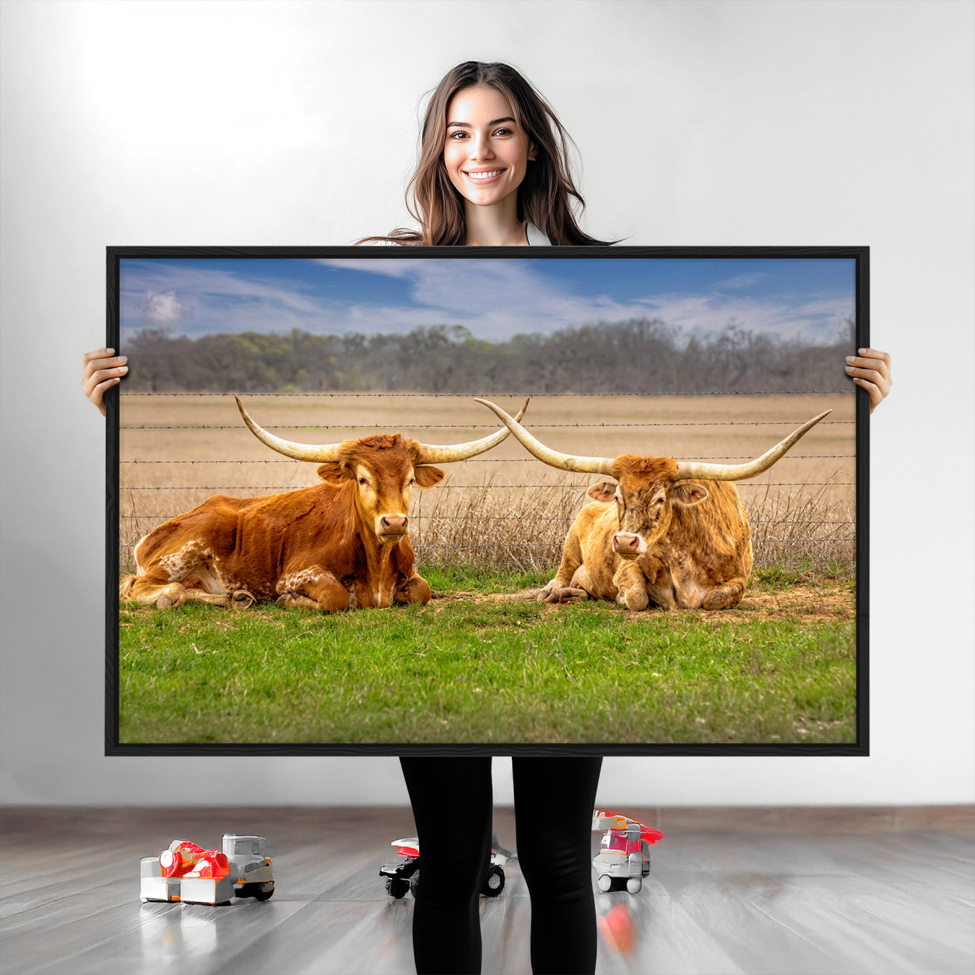 Two longhorn cows lying on green grass in front of a golden field and trees under a blue sky, perfect for rustic country wall decor.