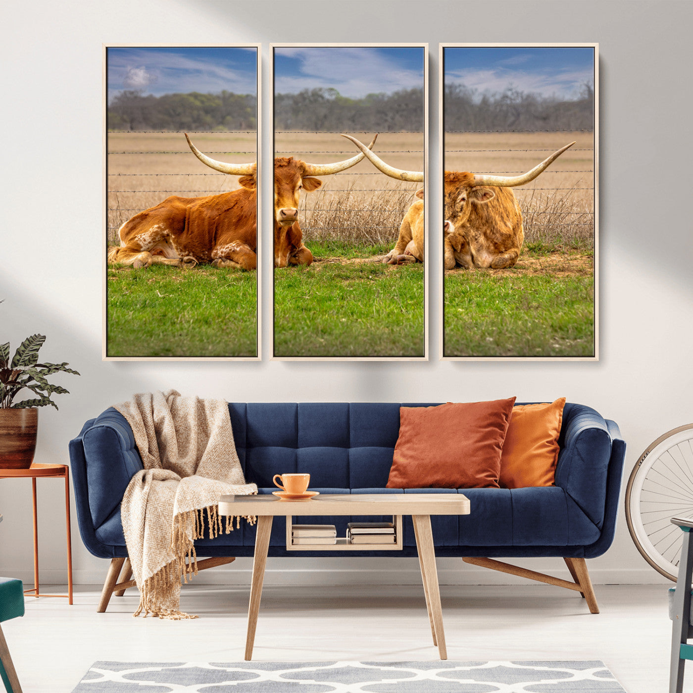 Two longhorn cows lying on green grass in front of a golden field and trees under a blue sky, perfect for rustic country wall decor.