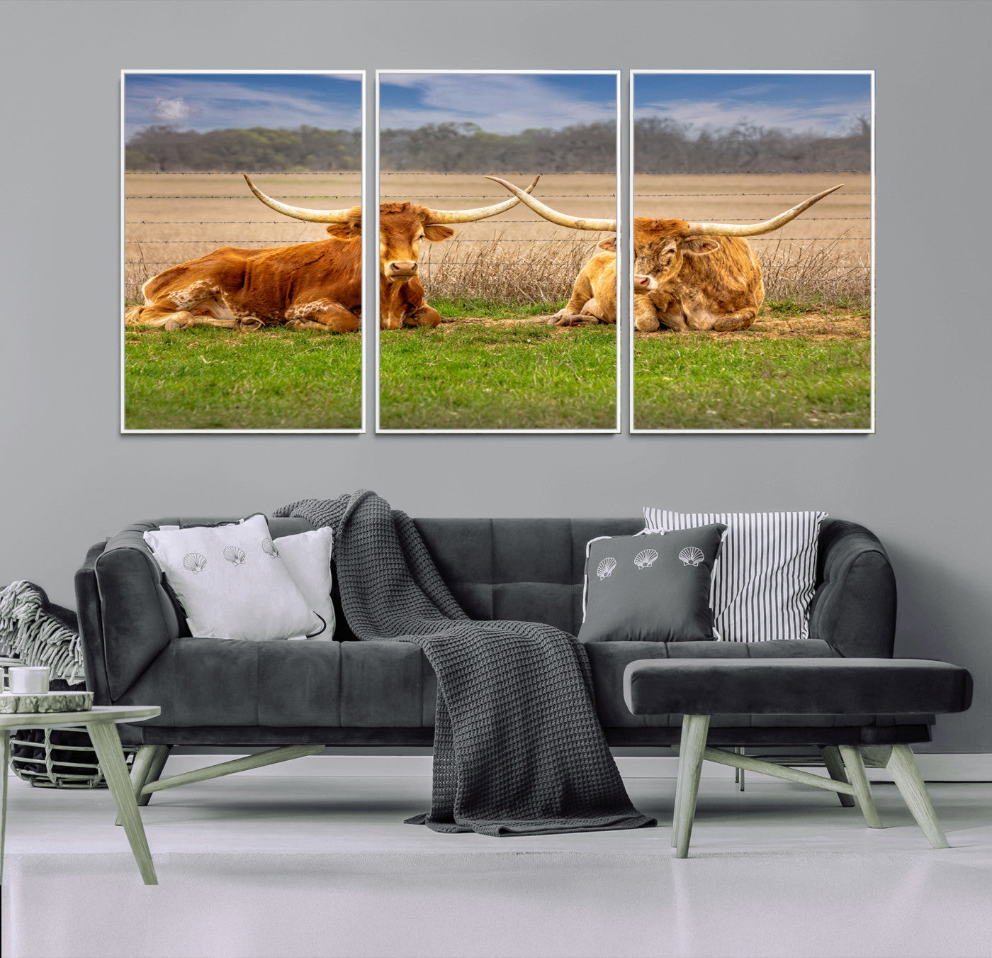 Two longhorn cows lying on green grass in front of a golden field and trees under a blue sky, perfect for rustic country wall decor.