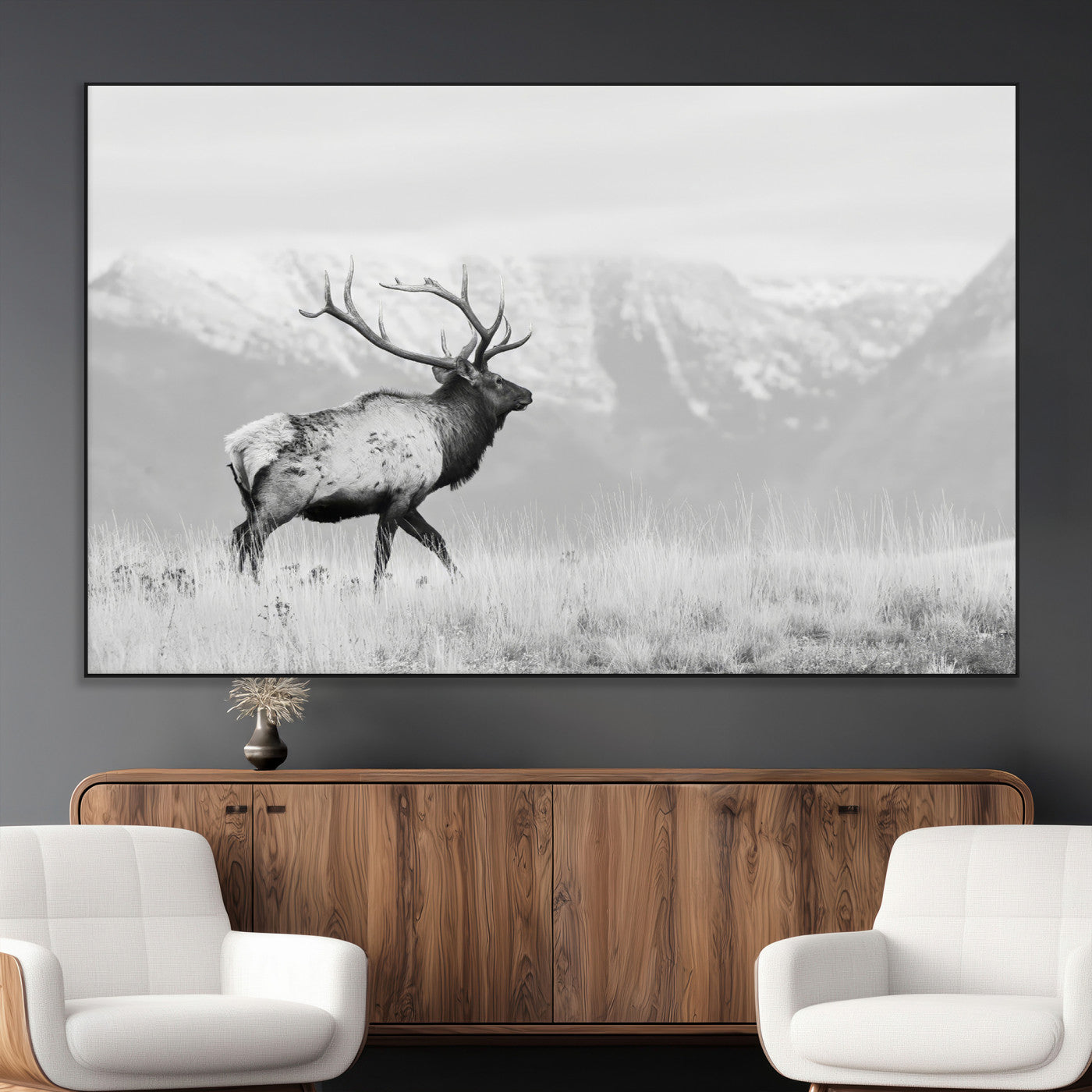 Black and white photograph of a majestic elk standing in a grassy field with snow-capped mountains in the background, creating a bold and rustic wilderness scene.