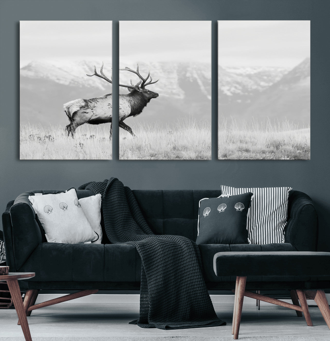 Black and white photograph of a majestic elk standing in a grassy field with snow-capped mountains in the background, creating a bold and rustic wilderness scene.