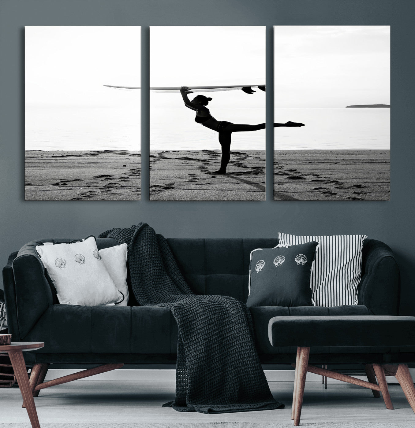 A woman balances a surfboard on her head while striking a yoga pose on the beach, silhouetted against a calm ocean horizon.