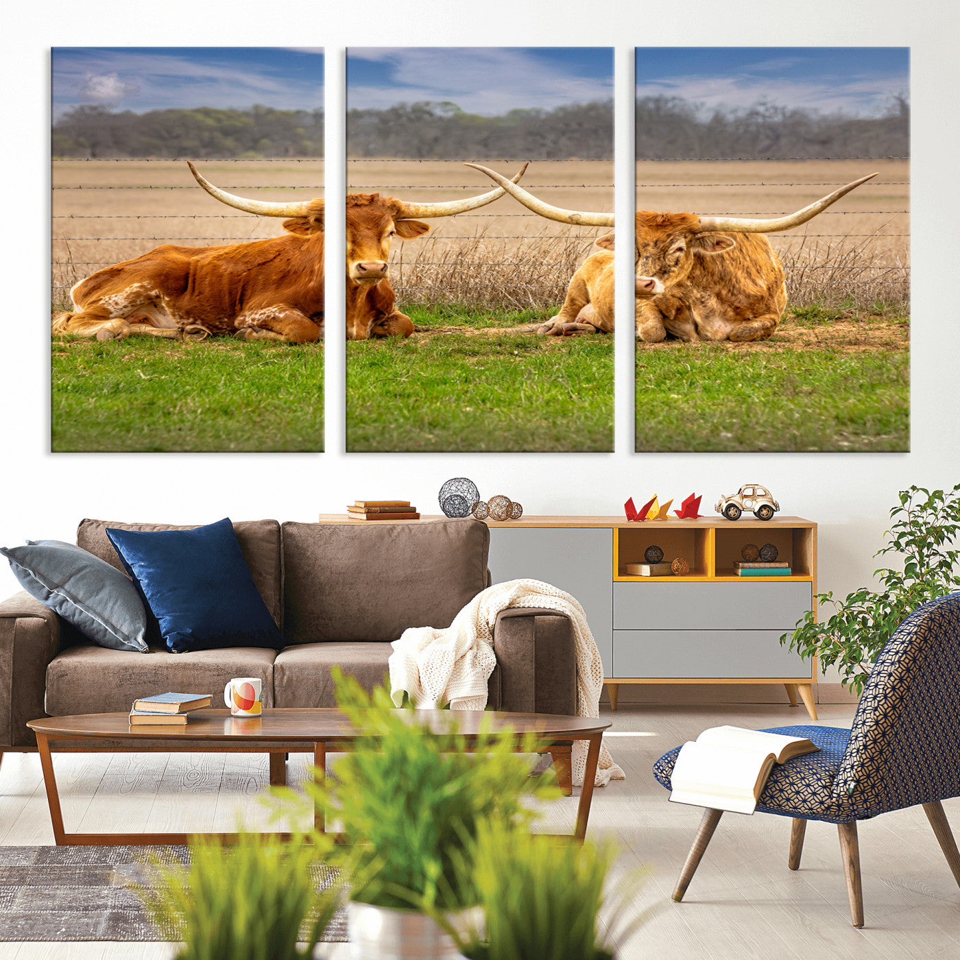 Two longhorn cows lying on green grass in front of a golden field and trees under a blue sky, perfect for rustic country wall decor.