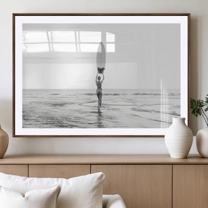 Black and white image of a female surfer holding a surfboard above her head while walking toward the ocean, creating a minimalist and bold coastal aesthetic.