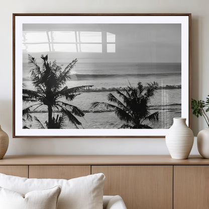Black and white view of ocean waves crashing on a tropical coastline, framed by silhouetted palm trees under a vast, open sky.