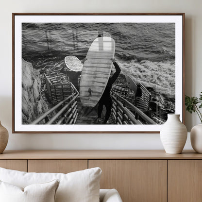 Black and white photo of surfers carrying boards down a rugged cliffside stairway, capturing a bold and adventurous coastal scene.