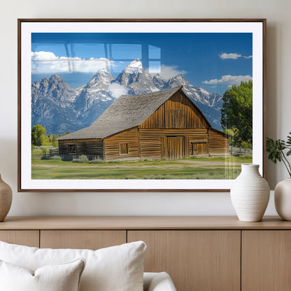 A rustic wooden barn sits in front of towering mountain peaks under a clear blue sky, perfect for farmhouse or cabin style interiors.