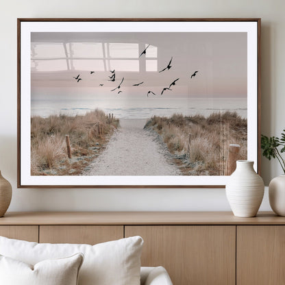 A peaceful coastal pathway leading to the ocean, with birds flying across a soft pastel sky, creating a calm and scenic beach atmosphere.