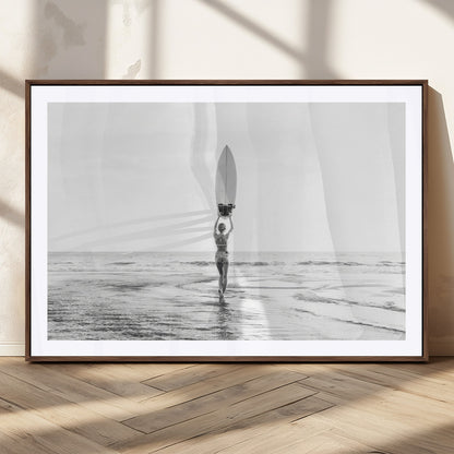Black and white image of a female surfer holding a surfboard above her head while walking toward the ocean, creating a minimalist and bold coastal aesthetic.