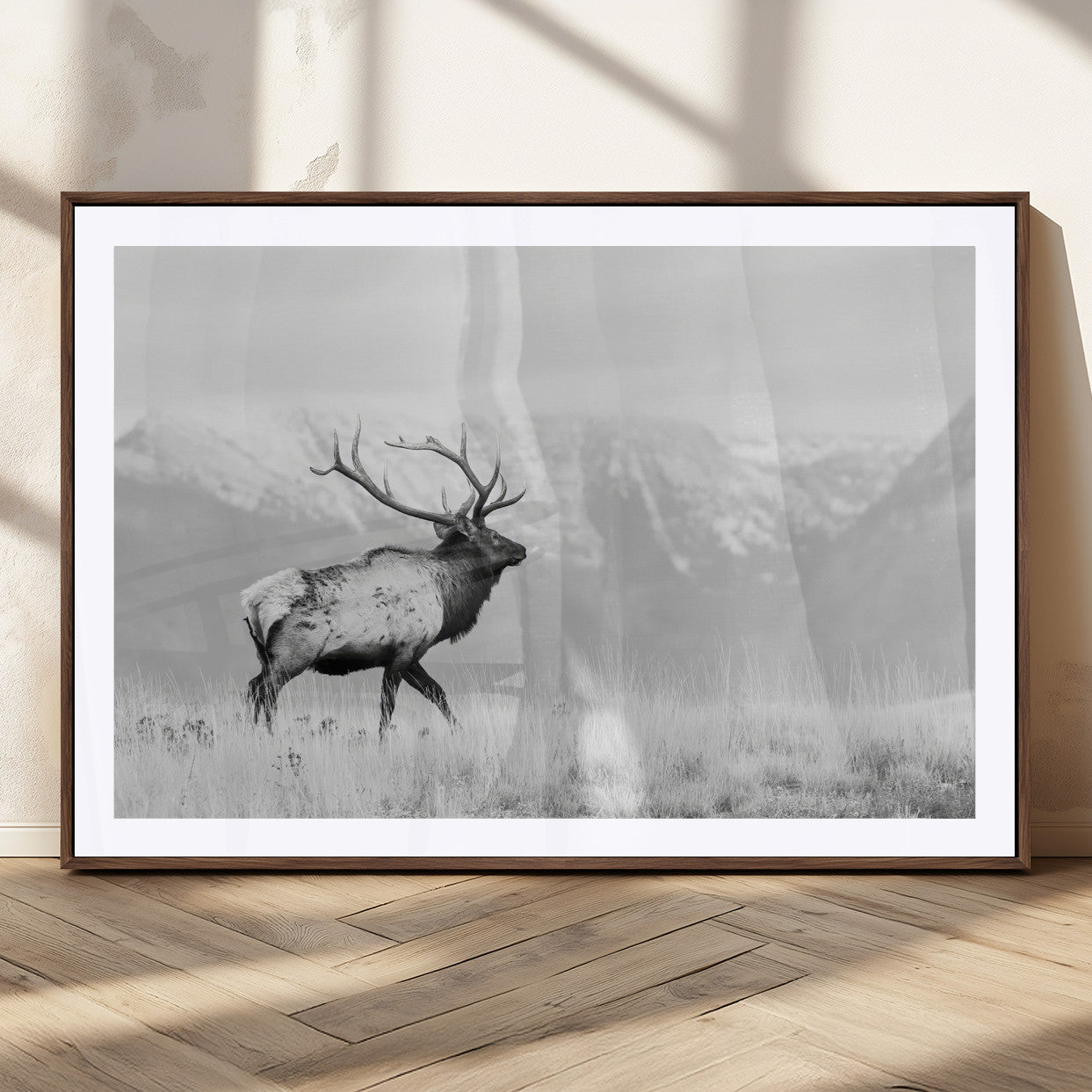 Black and white photograph of a majestic elk standing in a grassy field with snow-capped mountains in the background, creating a bold and rustic wilderness scene.