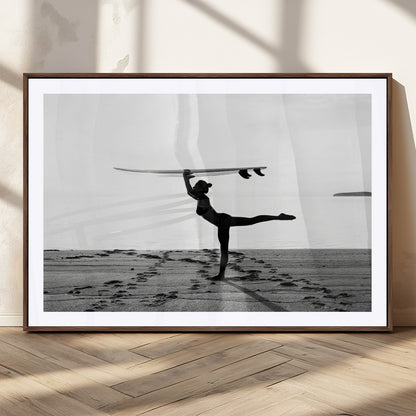 A woman balances a surfboard on her head while striking a yoga pose on the beach, silhouetted against a calm ocean horizon.