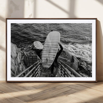 Black and white photo of surfers carrying boards down a rugged cliffside stairway, capturing a bold and adventurous coastal scene.