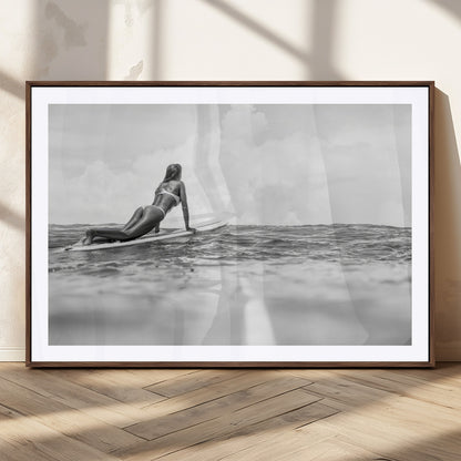 A black and white ocean scene featuring a surfer girl waiting on her board, gazing at the horizon, capturing a tranquil and adventurous coastal moment.