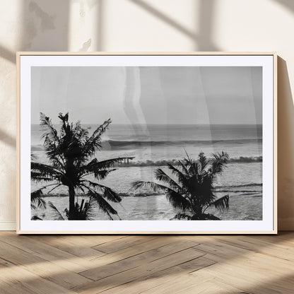 Black and white view of ocean waves crashing on a tropical coastline, framed by silhouetted palm trees under a vast, open sky.