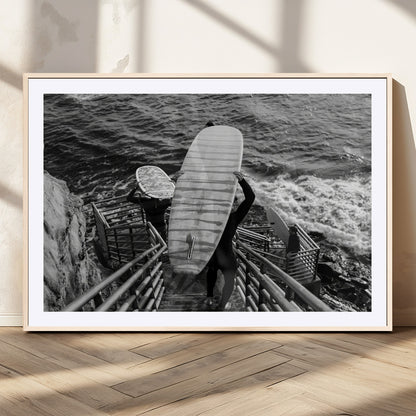 Black and white photo of surfers carrying boards down a rugged cliffside stairway, capturing a bold and adventurous coastal scene.
