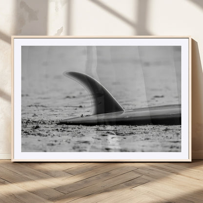 A black and white close-up of a surfboard fin resting on the sandy shore, highlighting minimalist beach aesthetics and classic surf culture.