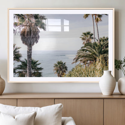 Scenic coastal view with tall palm trees overlooking a calm ocean, soft blue waves, and surfers in the distance, creating a peaceful tropical vibe.