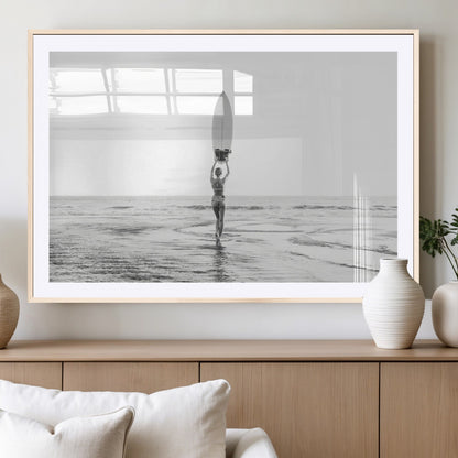 Black and white image of a female surfer holding a surfboard above her head while walking toward the ocean, creating a minimalist and bold coastal aesthetic.