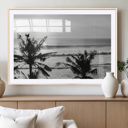 Black and white view of ocean waves crashing on a tropical coastline, framed by silhouetted palm trees under a vast, open sky.
