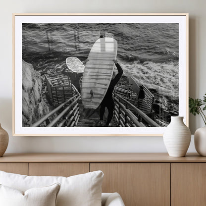 Black and white photo of surfers carrying boards down a rugged cliffside stairway, capturing a bold and adventurous coastal scene.