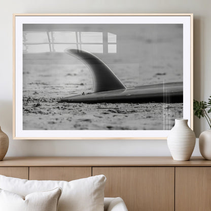 A black and white close-up of a surfboard fin resting on the sandy shore, highlighting minimalist beach aesthetics and classic surf culture.