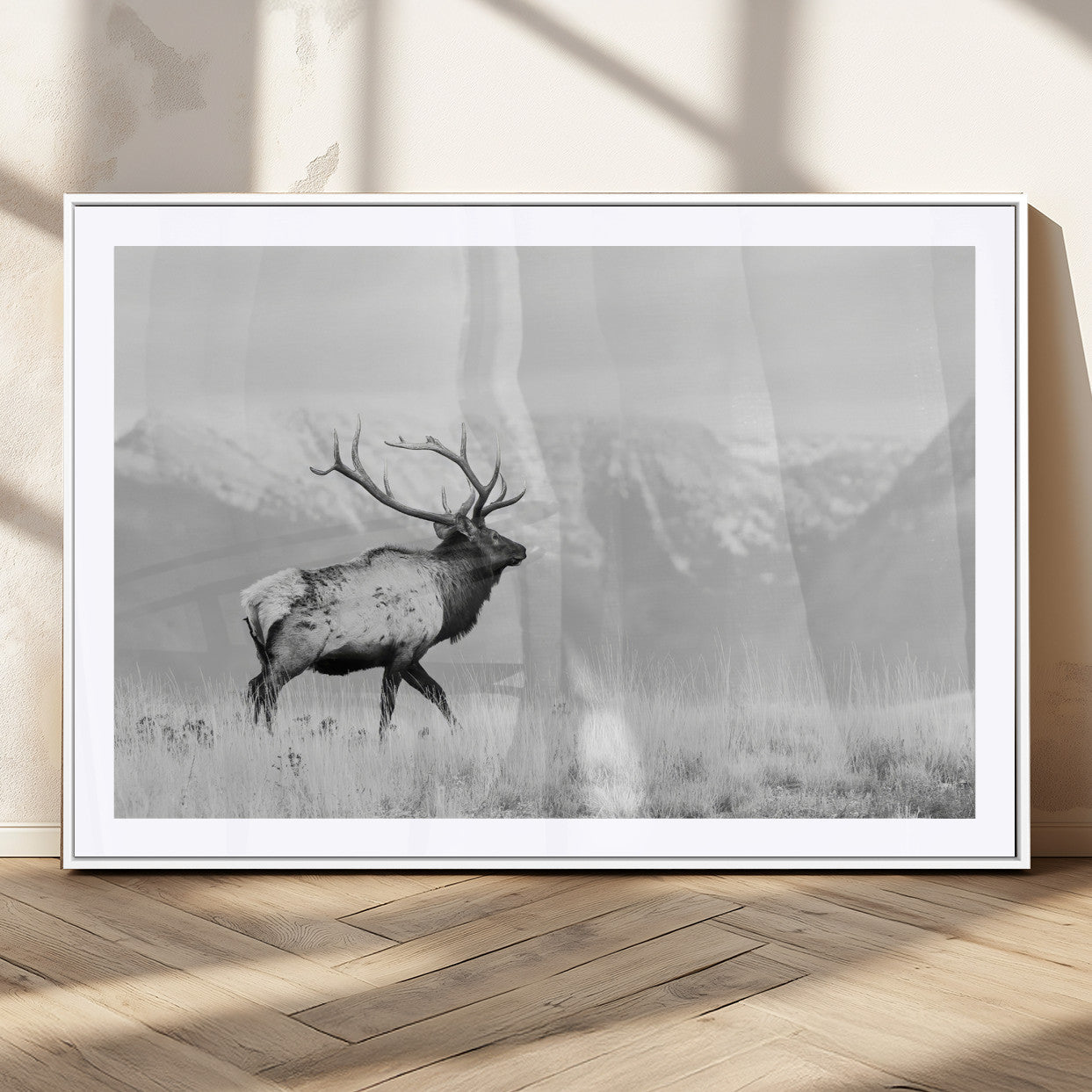 Black and white photograph of a majestic elk standing in a grassy field with snow-capped mountains in the background, creating a bold and rustic wilderness scene.