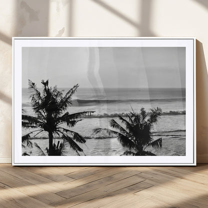 Black and white view of ocean waves crashing on a tropical coastline, framed by silhouetted palm trees under a vast, open sky.