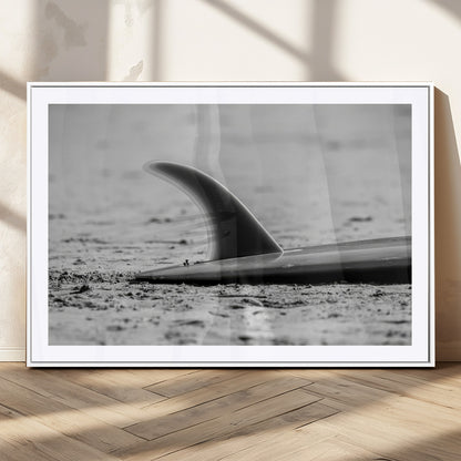 A black and white close-up of a surfboard fin resting on the sandy shore, highlighting minimalist beach aesthetics and classic surf culture.