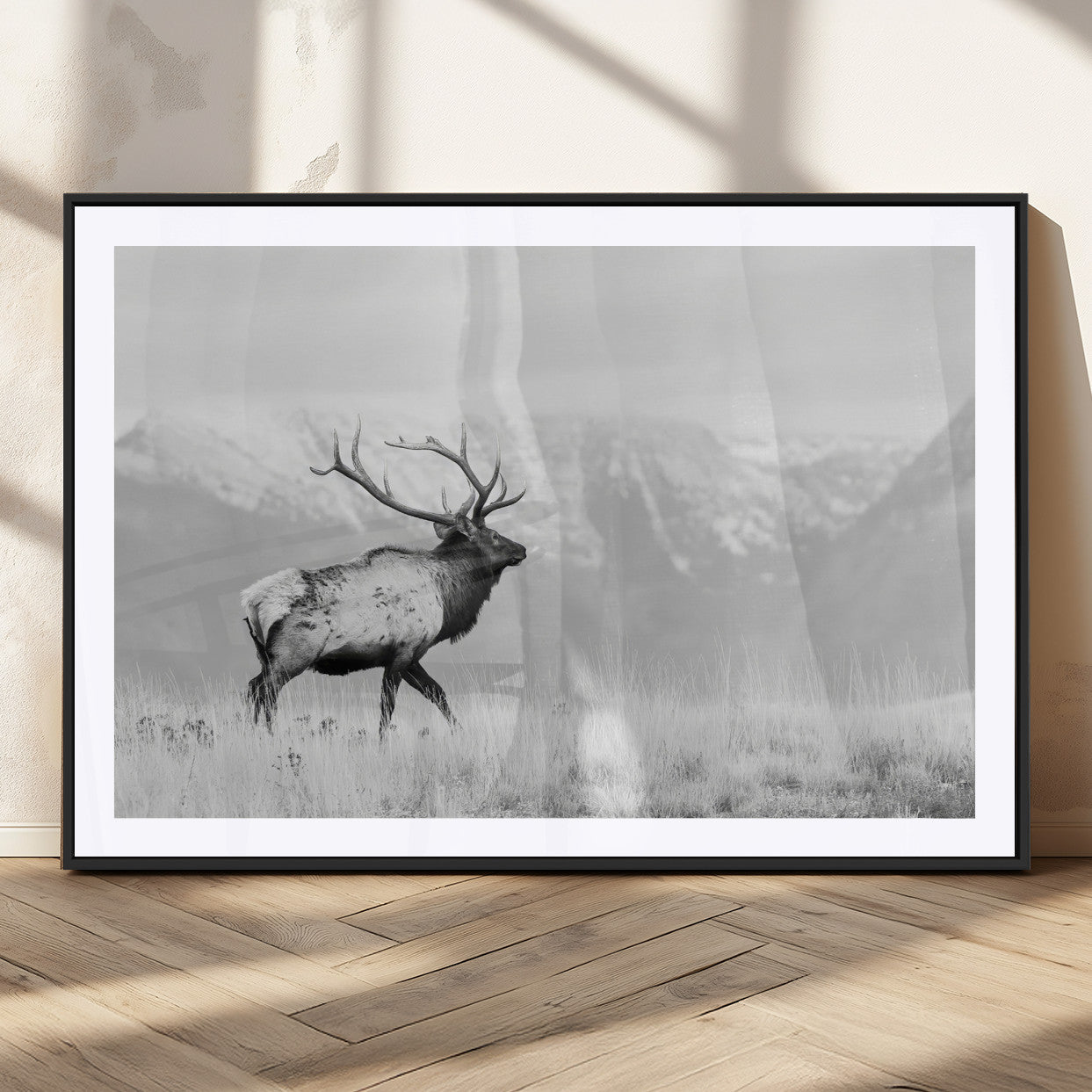 Black and white photograph of a majestic elk standing in a grassy field with snow-capped mountains in the background, creating a bold and rustic wilderness scene.