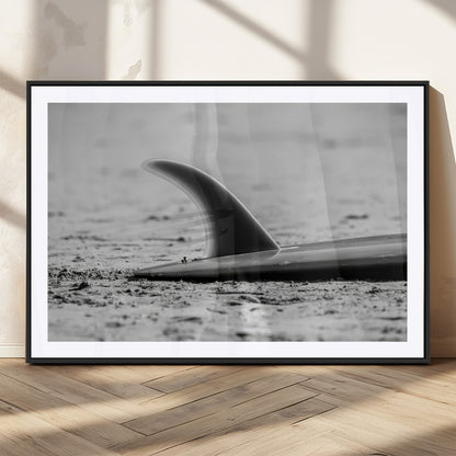 A black and white close-up of a surfboard fin resting on the sandy shore, highlighting minimalist beach aesthetics and classic surf culture.
