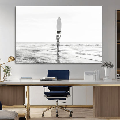 Black and white image of a female surfer holding a surfboard above her head while walking toward the ocean, creating a minimalist and bold coastal aesthetic.