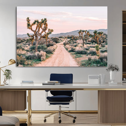 A dusty road winding through Joshua Tree National Park, framed by iconic twisted trees and warm desert tones under a soft pastel sky.