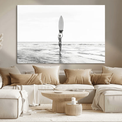 Black and white image of a female surfer holding a surfboard above her head while walking toward the ocean, creating a minimalist and bold coastal aesthetic.