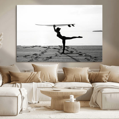 A woman balances a surfboard on her head while striking a yoga pose on the beach, silhouetted against a calm ocean horizon.