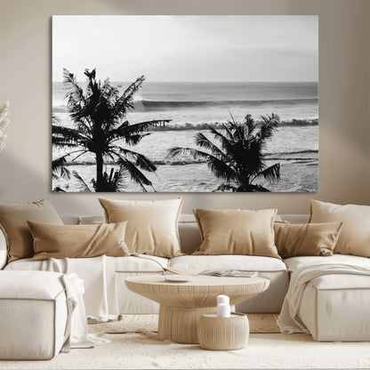 Black and white view of ocean waves crashing on a tropical coastline, framed by silhouetted palm trees under a vast, open sky.