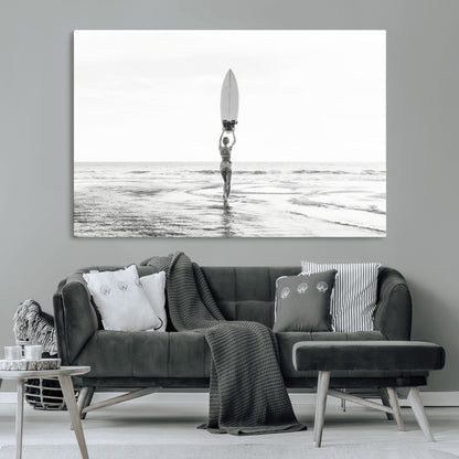 Black and white image of a female surfer holding a surfboard above her head while walking toward the ocean, creating a minimalist and bold coastal aesthetic.