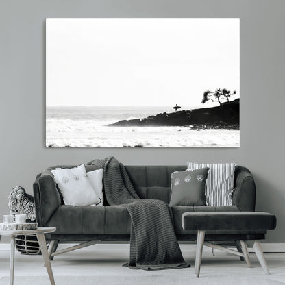 Black and white photo of a surfer standing on rocky cliffs, gazing at the waves, embodying the spirit of adventure and solitude.