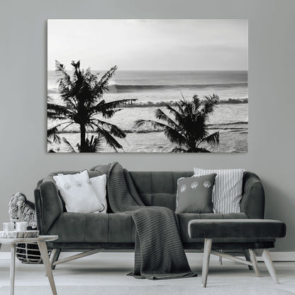 Black and white view of ocean waves crashing on a tropical coastline, framed by silhouetted palm trees under a vast, open sky.