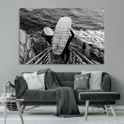 Black and white photo of surfers carrying boards down a rugged cliffside stairway, capturing a bold and adventurous coastal scene.