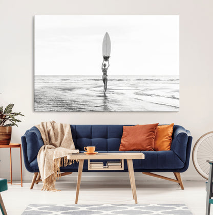 Black and white image of a female surfer holding a surfboard above her head while walking toward the ocean, creating a minimalist and bold coastal aesthetic.