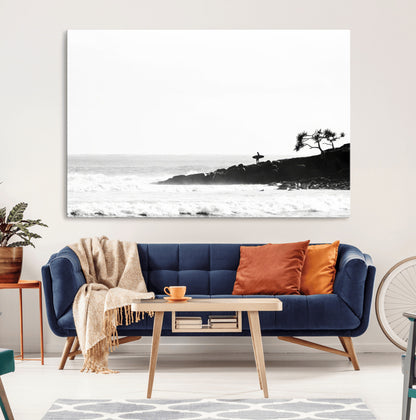 Black and white photo of a surfer standing on rocky cliffs, gazing at the waves, embodying the spirit of adventure and solitude.