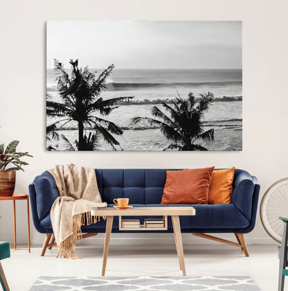 Black and white view of ocean waves crashing on a tropical coastline, framed by silhouetted palm trees under a vast, open sky.