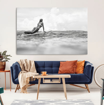 A black and white ocean scene featuring a surfer girl waiting on her board, gazing at the horizon, capturing a tranquil and adventurous coastal moment.