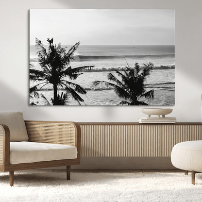 Black and white view of ocean waves crashing on a tropical coastline, framed by silhouetted palm trees under a vast, open sky.