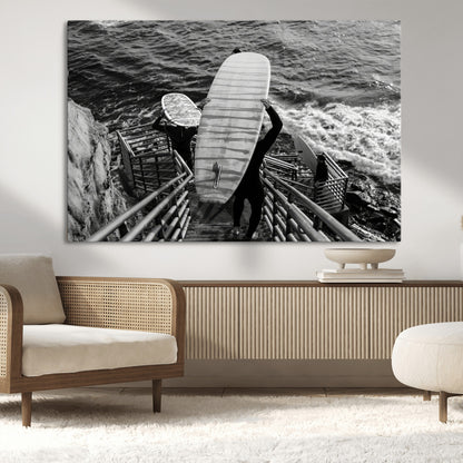 Black and white photo of surfers carrying boards down a rugged cliffside stairway, capturing a bold and adventurous coastal scene.