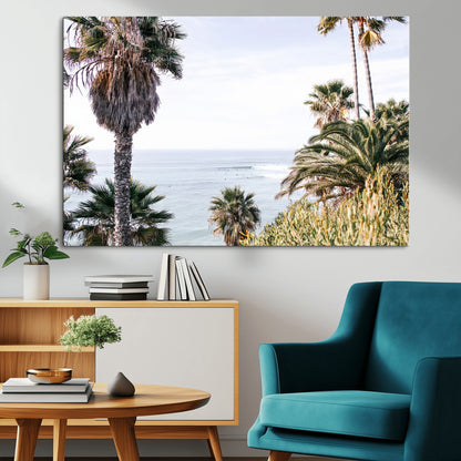 Scenic coastal view with tall palm trees overlooking a calm ocean, soft blue waves, and surfers in the distance, creating a peaceful tropical vibe.